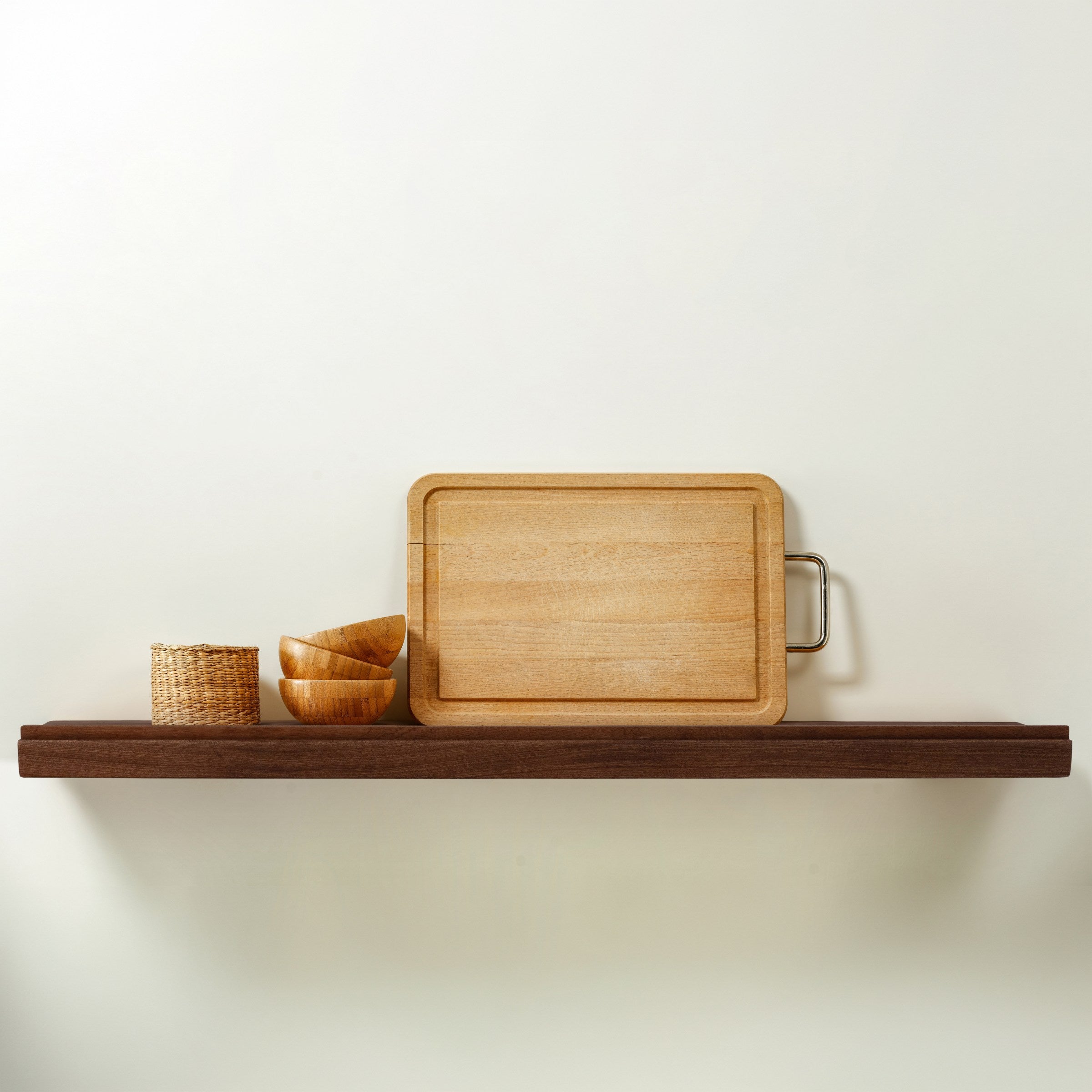A rectangular light wood cutting board with a metal handle resting on a dark wooden floating shelf with a lip on the front edge, alongside a small woven basket and stacked wooden bowls, against a plain off-white wall.